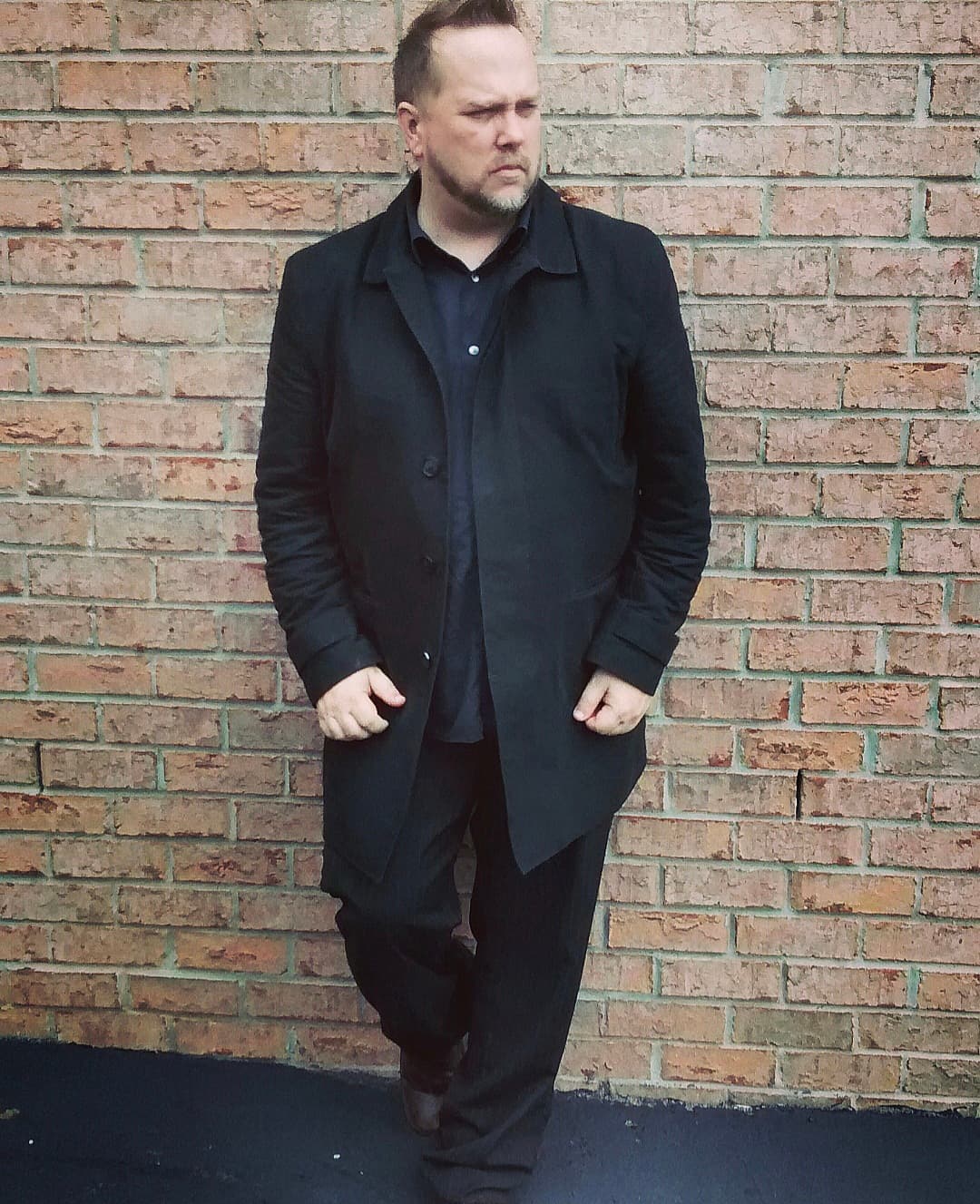 Full-body portrait of Kene Bell dressed in black against a brick wall