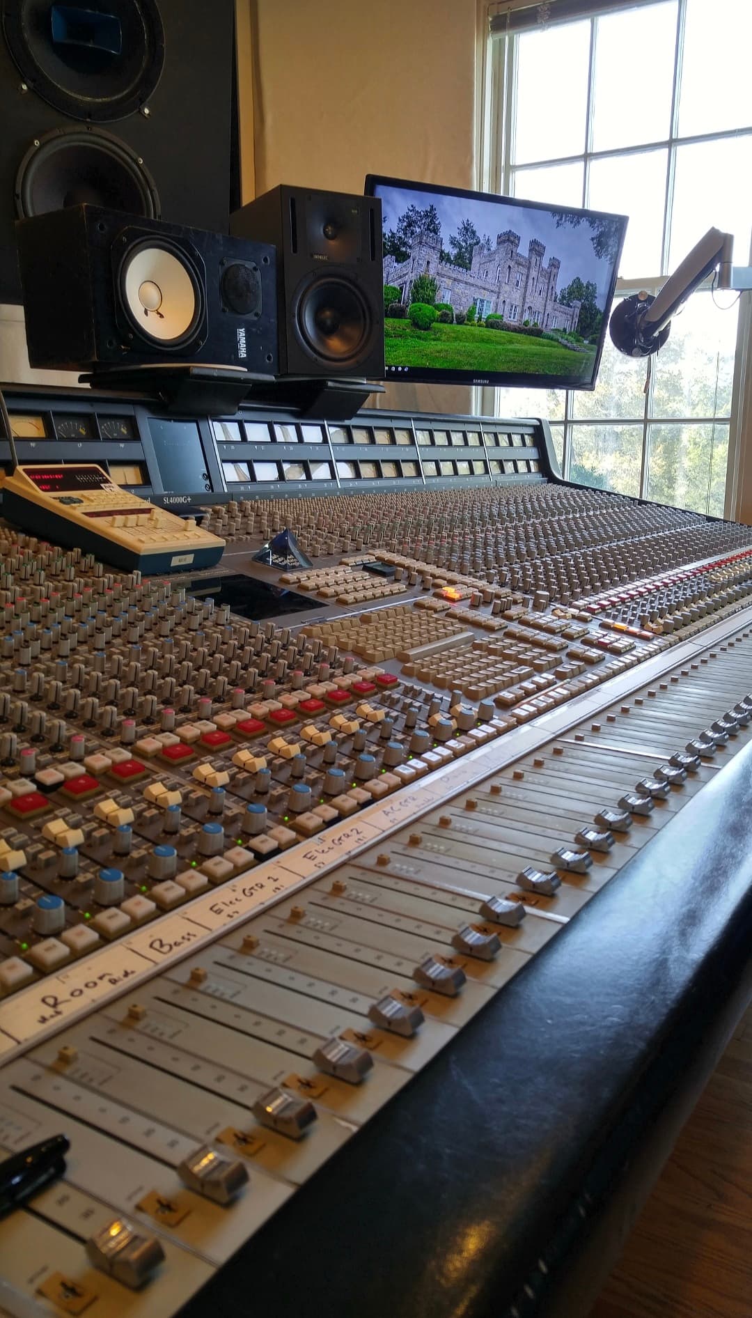 Daylight view across a large analog mixing console with studio monitors and a screen positioned in front of a window