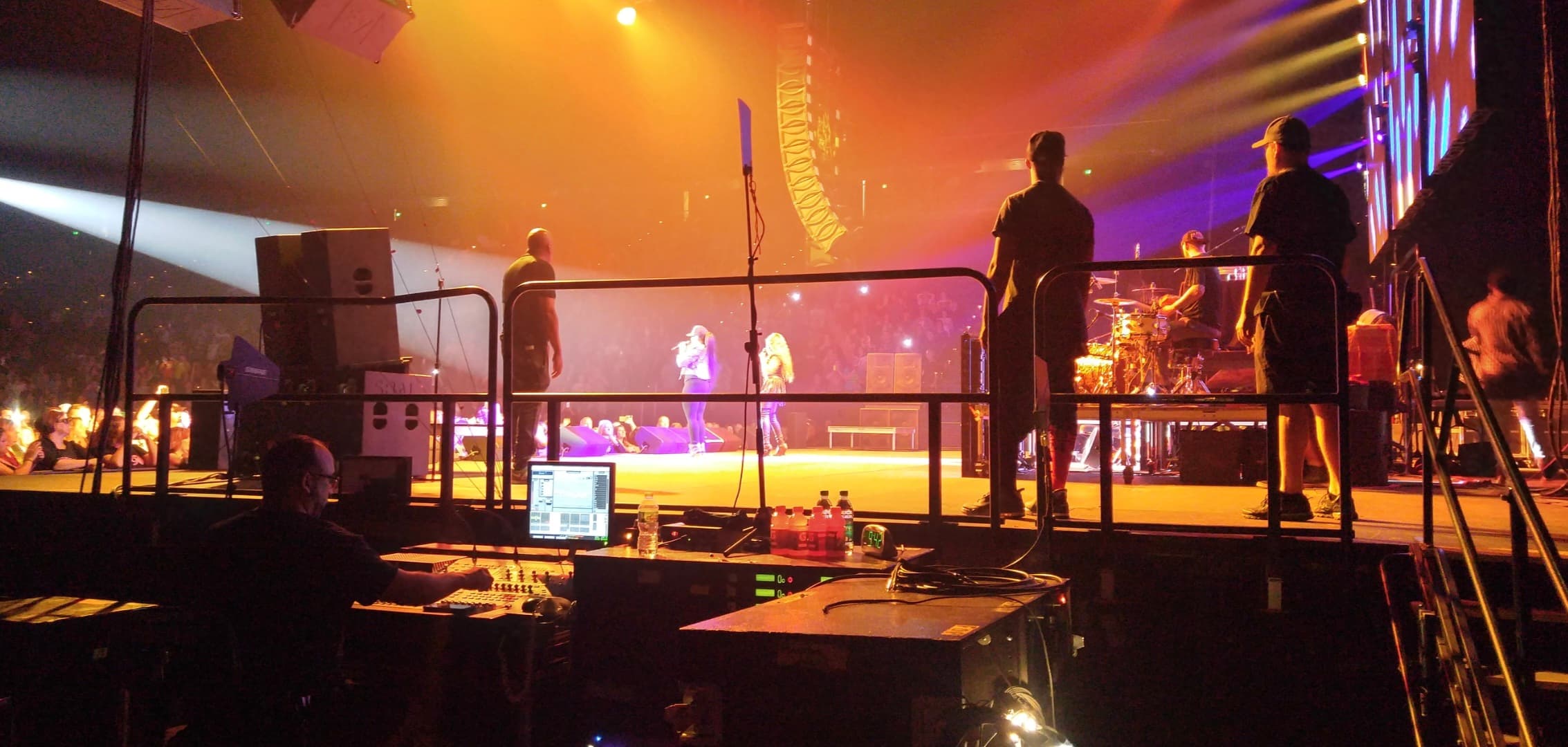 Side-stage view across a live production position, performers, musicians, lighting, and audience